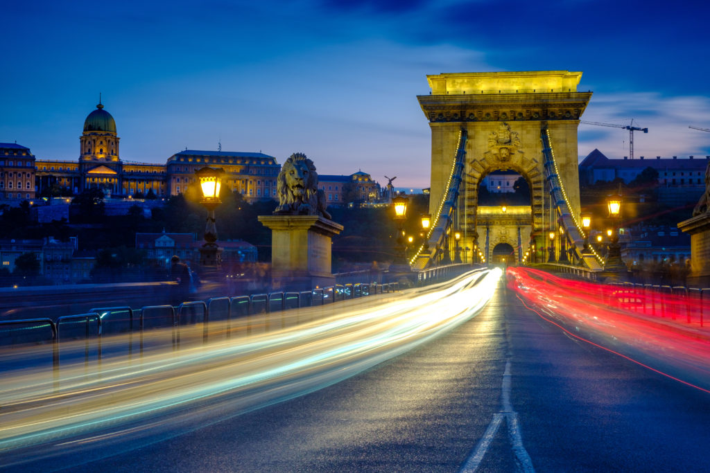 Plan crépusculaire de la circulation et des sentiers de lumière traversant le Pont des Chaînes à Budapest.  Les lumières de la ville sont allumées et le château de Buda est en arrière-plan